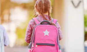 Dziewczynka w różowej koszuli w kratę z dwoma warkoczami nosi jasnoróżowy plecak z szarą gwiazdką.
