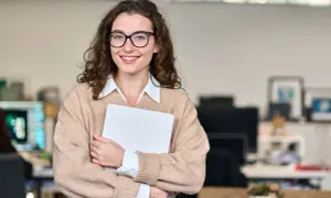 Uśmiechnięta kobieta w okularach trzyma laptopa. Jej strój i pewność siebie sugerują, że doskonale odnajduje się w pracy związanej z obsługą klienta, kod zawodu.