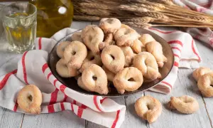 Piatto pieno di taralli dolci al vino bianco, cosparsi di zucchero. Accanto, un bicchiere di vino bianco e una bottiglia d'olio.