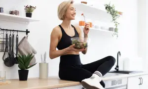Une femme souriante en tenue de sport mange une salade fraîche. Elle savoure son repas sain dans une cuisine lumineuse.