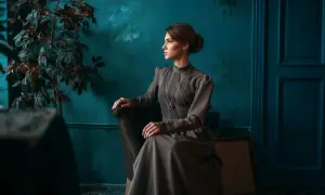 Elegancka kobieta w sukni w stylu dark academia siedzi w fotelu, wpatrzona w dal. W tle ciemnozielona ściana i rośliny.
