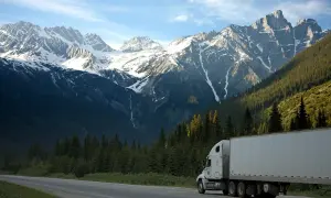 Un camion Scania V8 blanc roule sur une route de montagne bordée de forêts denses, sous un ciel bleu et des sommets enneigés.