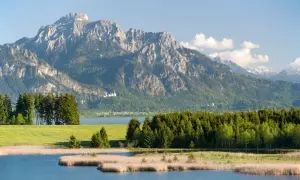 Majestätischer berg mit see und einer kleinen Burg im Hintergrund. Grüne Wiesen und Schilf am Ufer.