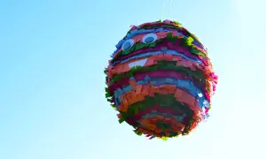 Piñata casera colorida con ojos saltones y boca sonriente, colgada contra un cielo azul claro.