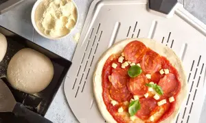 Pizzadeg recept: färdig pizzadeg med salami, ost och basilika på en pizzaspade. Bredvid ligger mer deg och en skål med mjöl.