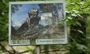 Tablica informacyjna w lesie przedstawia skalne formacje i korzenie drzew. Podpis "Prządka-Matka" sugeruje, skąd pochodzi nazwa rezerwatu prządki, nawiązując do legendy.