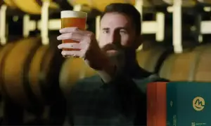 Uomo con barba esamina un bicchiere di birra dorata, con botti di legno sullo sfondo. Scatole di birre lager famose in primo piano.