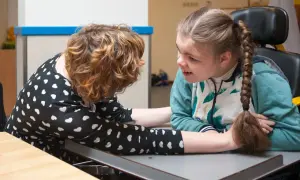 Dwie dziewczynki przy stole, jedna z warkoczem, druga pochylona. W tle widać elementy sali terapeutycznej, sugerujące opóźnienie rozwoju a autyzm.