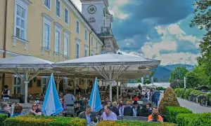 Menschen genießen die Tegernsee Feste unter Sonnenschirmen. Ein Gebäude mit Turmuhr und bayerische Fahnen im Hintergrund.