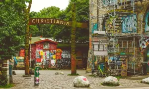Drewniany totem i tablica z napisem "CHRISTIANIA" wita w tej wolnej dzielnicy. Obok czerwony budynek z plakatami i rowery.