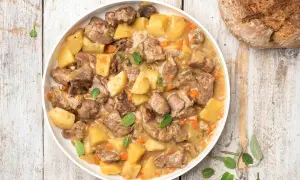 Un saporito agnello in umido con patate, carote e erbe aromatiche, servito in un piatto bianco su un tavolo di legno rustico.