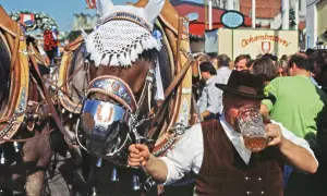 Prachtvolle Pferde mit Trachtenschmuck, ein Mann im Lederhosen-Look trinkt Bier. Ein typischer Hintergrund Oktoberfest-Stimmung.