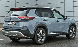 Szary Nissan X-Trail e-POWER z czarnym dachem i srebrnymi akcentami stoi na mokrym parkingu przed nowoczesnym budynkiem.