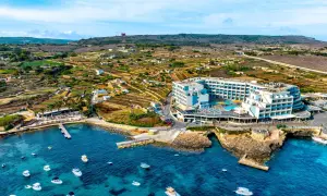 Malta last minute tanio: hotel z basenem nad turkusowym morzem, otoczony tarasami i łodziami. W tle malownicze, tarasowe pola i wzgórza.