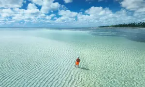 Człowiek w pomarańczowych spodenkach stoi w płytkiej, turkusowej wodzie. Zastanawia się, ile się leci na Zanzibar, by dotrzeć do tego raju.
