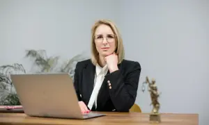 Kobieta w okularach i żakiecie siedzi przy biurku z laptopem i figurką Temidy.