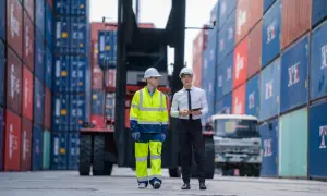 Dwóch mężczyzn w kaskach stoi między rzędami kontenerów w terminalu portowym.