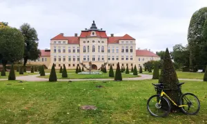 Pałac w Strykowie z żółtą ramą roweru na pierwszym planie, otoczony zadbanym parkiem z rzędami stożkowych drzew.