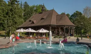 Wysowa Zdrój atrakcje: fontanna, drewniana karczma i zielony park. Ludzie odpoczywają przy stolikach pod parasolami.