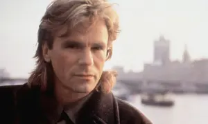 MacGyver obsada: Richard Dean Anderson w płaszczu, z charakterystyczną fryzurą, na tle zamglonego miasta.