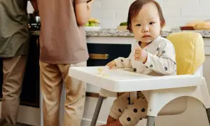 Małe dziecko siedzi w krzesełku dla dziecka, jedząc kawałki banana. W tle widać dwie osoby przygotowujące posiłek w kuchni.