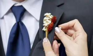 Dłoń przypina do marynarki mały bukiecik z suszonych kwiatów. To właśnie jest butonierka co to za ozdoba.