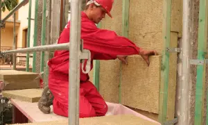 Budowlaniec w czerwonym stroju montuje wełnę mineralną na fasadzie. Stosuje metodę lekką suchą, dbając o izolację budynku.