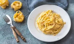 Un plat de fettuccine alfredo crémeux, saupoudré de poivre noir, avec des pâtes crues et une fourchette en bois à côté.