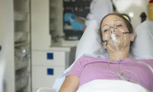 Młoda kobieta w masce tlenowej, leżąca w łóżku szpitalnym. Jej stan sugeruje chorobę popromienną, wymagającą intensywnej opieki medycznej.