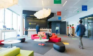 Un **open space** moderne chez Microsoft. Une femme travaille sur son ordinateur portable, entourée de canapés colorés. Des affiches "Imagine", "Achieve", "Innovate" flottent au plafond.