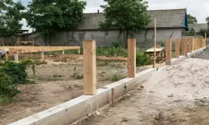 Solidny fundament pod ogrodzenie z betonowym murem i drewnianymi słupkami. W tle widać budowę domu.