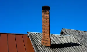 Ceglany komin na dachu, z którego wydobywa się sadza, na tle błękitnego nieba.