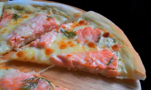 Kawałek pysznej pizzy z łososiem, roztopionym serem i koperkiem na drewnianej desce.