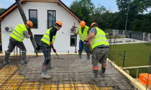 Budowlańcy w kamizelkach i kaskach, przestrzegając zasady bhp, wylewają beton. Jeden kieruje rurę, drugi grabi, trzeci wyrównuje.