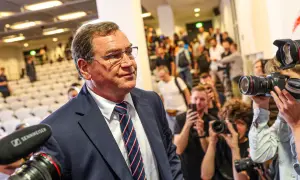 Mężczyzna w garniturze, otoczony przez fotoreporterów z aparatami, przypomina jednego z kapitanów reprezentacji Polski, gotowego na ważne wydarzenie.