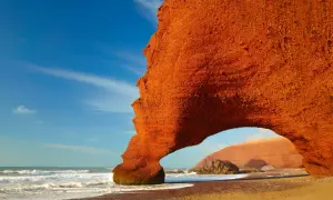 Pomarańczowe skały na plaży w Maroku, fale oceanu i błękitne niebo. Niesamowite widoki, a ceny w Maroku zachęcają do podróży.