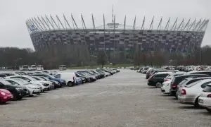Duży parking przed Stadionem Narodowym w Warszawie. Widać rzędy samochodów i autobusów na błoniach stadionu.