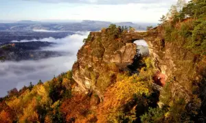 Majestätische Felsformation Pravcicka brana mit einem natürlichen Torbogen, umgeben von herbstlichem Laub und Nebel im Tal.