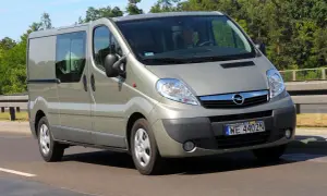 Srebrny Opel Vivaro 2.0 CDTI z przyciemnianymi szybami jedzie po drodze, otoczony zielenią drzew.