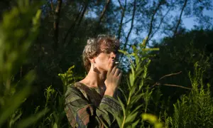 Młoda osoba w okularach i zielonej tunice z papierosem w dłoni, otoczona bujną roślinnością. Obraz w stylu polscy fotografowie współcześni.
