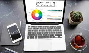 Laptop z kołem kolorów i paletą barw, obok telefon, notes i kaktus. Idealne narzędzia do pracy nad projektami z wykorzystaniem rgb druk.