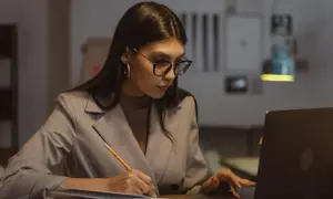 Kobieta w okularach pracuje przy laptopie, notując w zeszycie. Zastanawia się nad limitem nadgodzin w roku, by zachować równowagę.