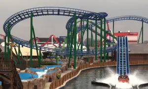 Water coaster zjeżdża z dużej wysokości, tworząc fontannę wody. W tle widać inne atrakcje parku rozrywki.