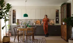 En kvinna i orange jumpsuit lutar sig mot köksbänken i ett kök med fransk inredningsstil. Ett runt träbord med stolar står framför köksöarna.