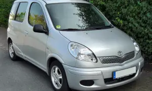 Srebrny Toyota Yaris Verso zaparkowany przy żywopłocie.