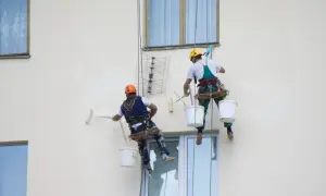 Deux peintres suspendus par des cordes peignent un mur extérieur. L'un utilise un rouleau, l'autre est prêt à commencer.