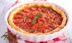 Une délicieuse tarte à la tomate, garnie de fines herbes et d'une branche de romarin frais, repose dans un plat blanc sur un torchon à carreaux rouges.