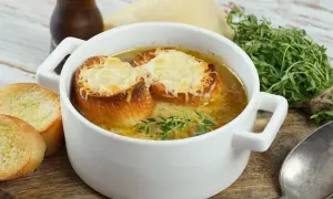 Aksamitna zupa cebulowa przepis, podana w białej miseczce, z grzankami zapieczonymi z serem i gałązką tymianku. Obok kromki chleba.