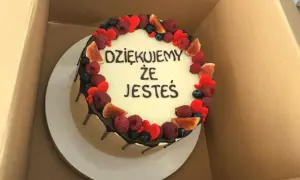Okrągły tort z białym kremem, ozdobiony owocami i serduszkami. Krótki napis na tort urodzinowy: "DZIĘKUJEMY ŻE JESTEŚ".