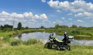 Motocyklista w kasku siedzi na motocyklu obok rzeki, na tle zielonej łąki i błękitnego nieba z chmurami.